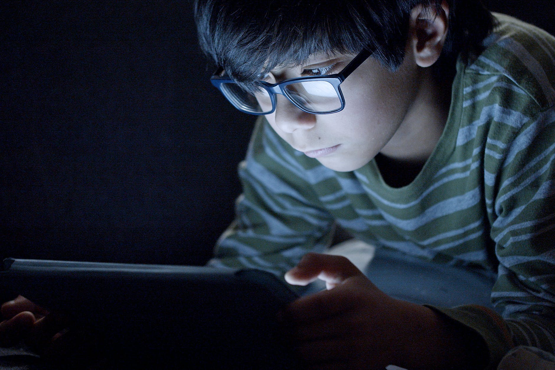 boy wearing eyeglasExcessive screen time causing dry eye problems in kidsses using a tablet in the dark room